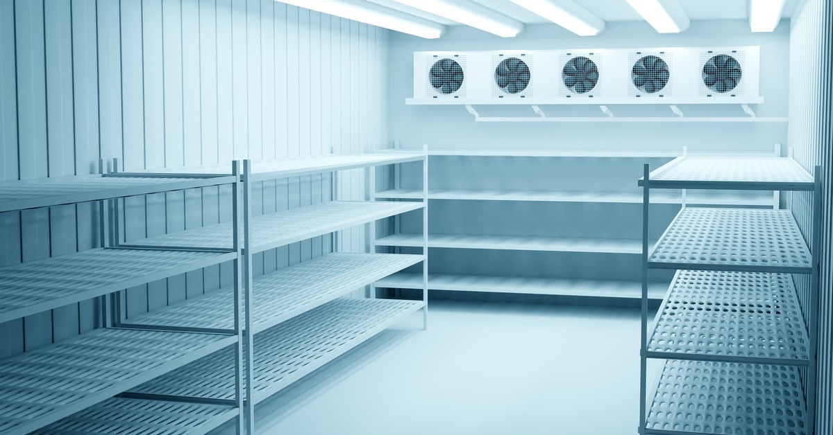 The inside of a walk-in cooler. The ceiling has several fluorescent light bulbs, and the walls are lined with shelf units.