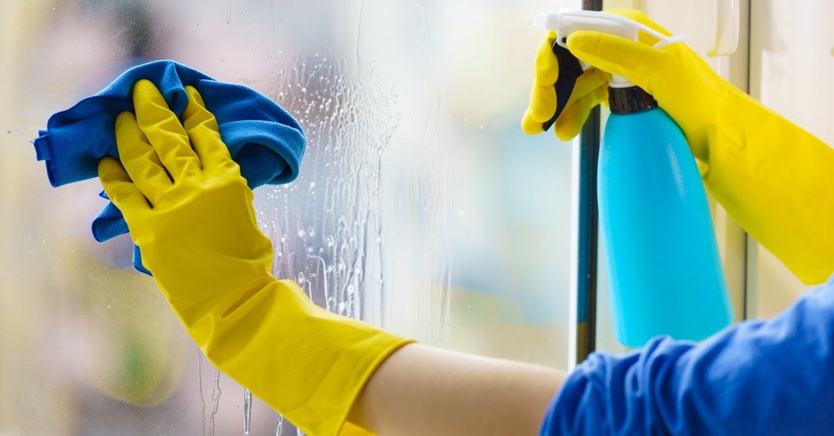 A pair of hands wearing yellow rubber gloves. They're spraying a glass panel with cleaner and wiping it with a blue cloth.